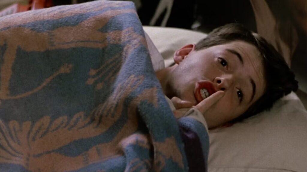 A young man lying in bed under a patterned blanket, holding a finger to his lips in a shushing gesture.