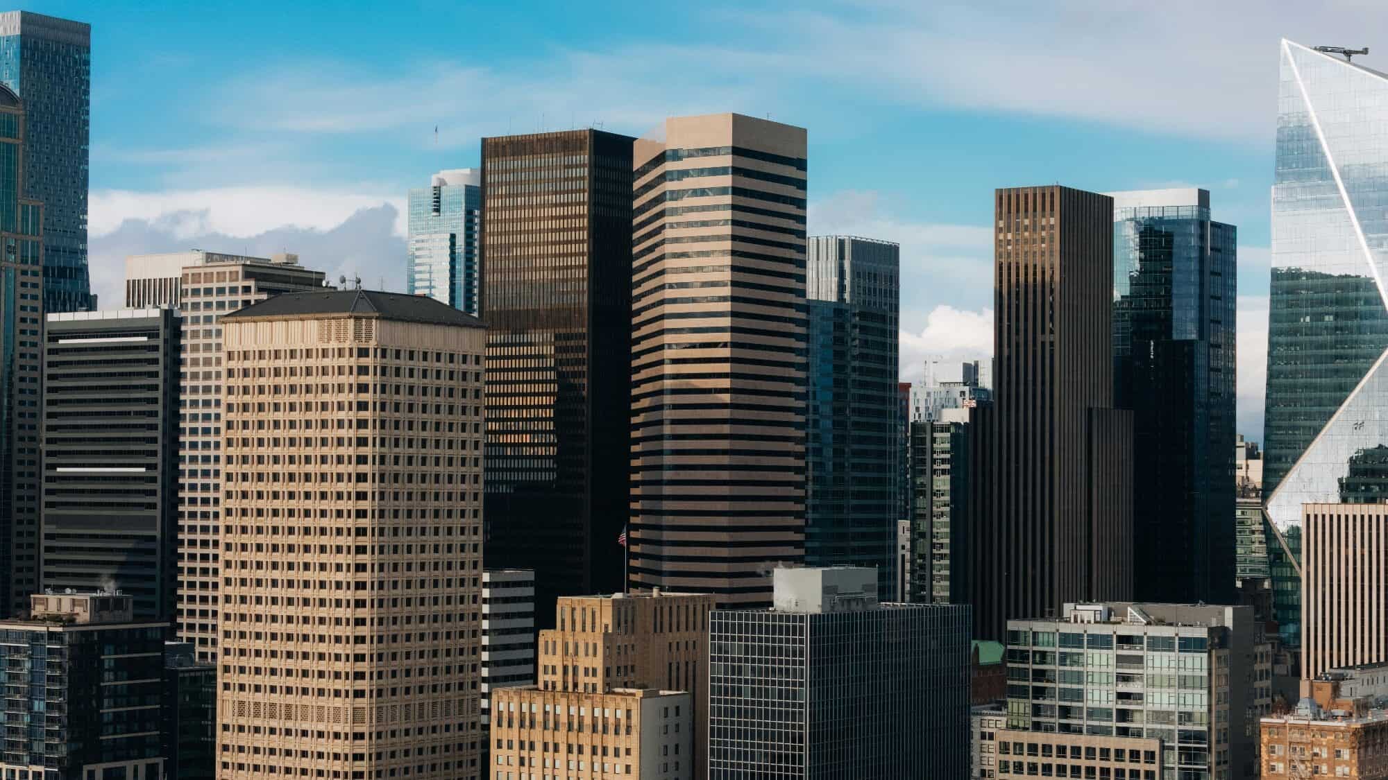 josh-hild-L0Jrd-c-I0o-unsplash-16-9-2000x1125 A cluster of modern high-rise office buildings with glass and concrete facades against a blue sky with some clouds.