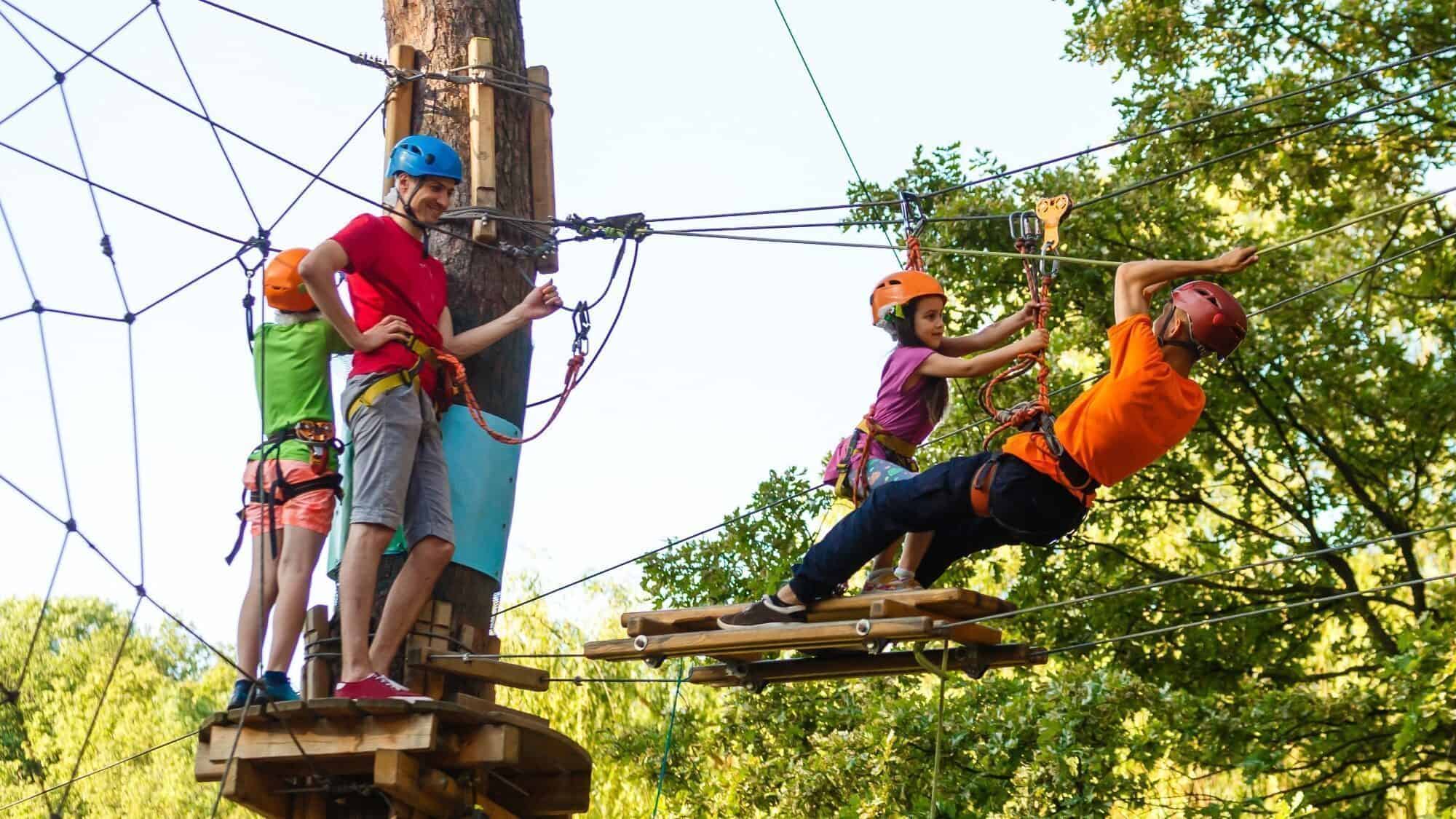 Four people wearing helmets and harnesses navigate a ropes course outdoors, with two standing on a platform and two balancing on a suspended wooden bridge.