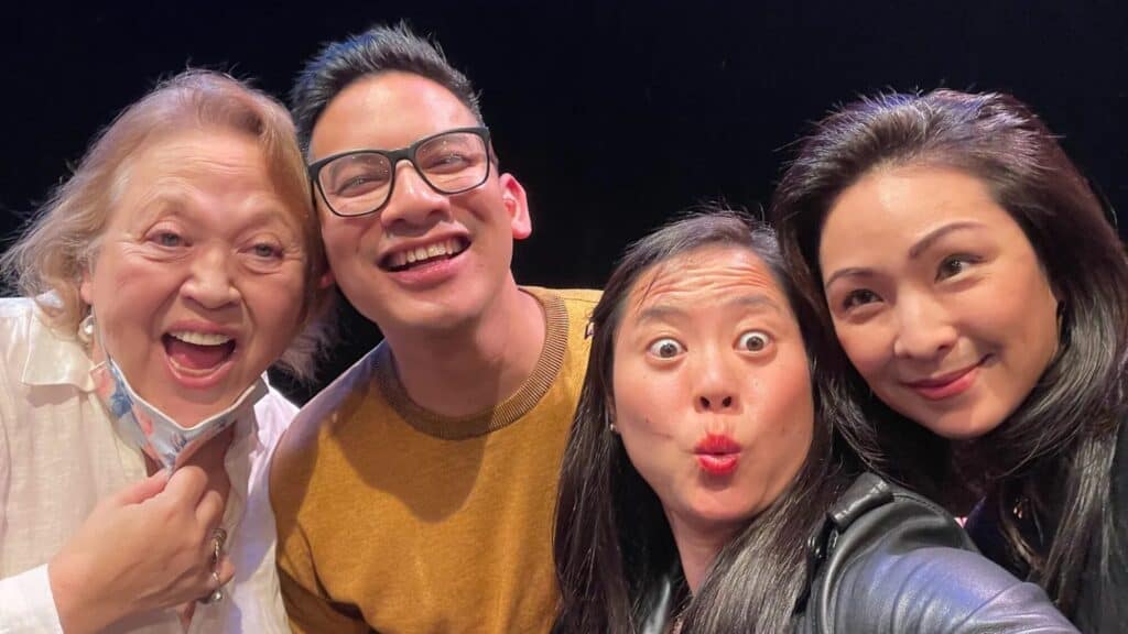 Four people smile and pose for a selfie against a dark background at Theatre Off Jackson; one, the Interim Executive Director, wears glasses while another makes a puckered face.