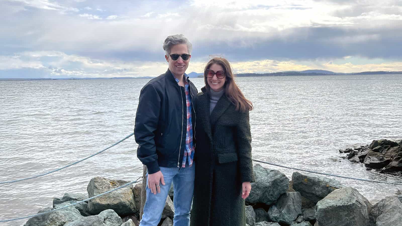 A man and a woman stand on rocks by the water, both wearing sunglasses and coats, with a cloudy sky and calm waves in the background.