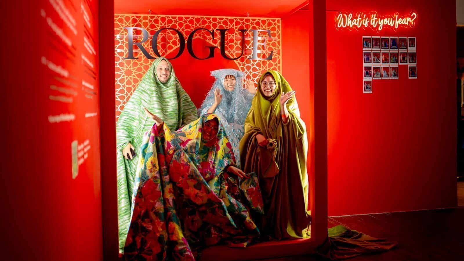 Four people draped in colorful, patterned fabrics pose in front of a red wall with "ROGUE" and a neon "What is it you fear?"—a vivid scene that could easily be found at the Seattle Art Fair, celebrating innovative Seattle art.