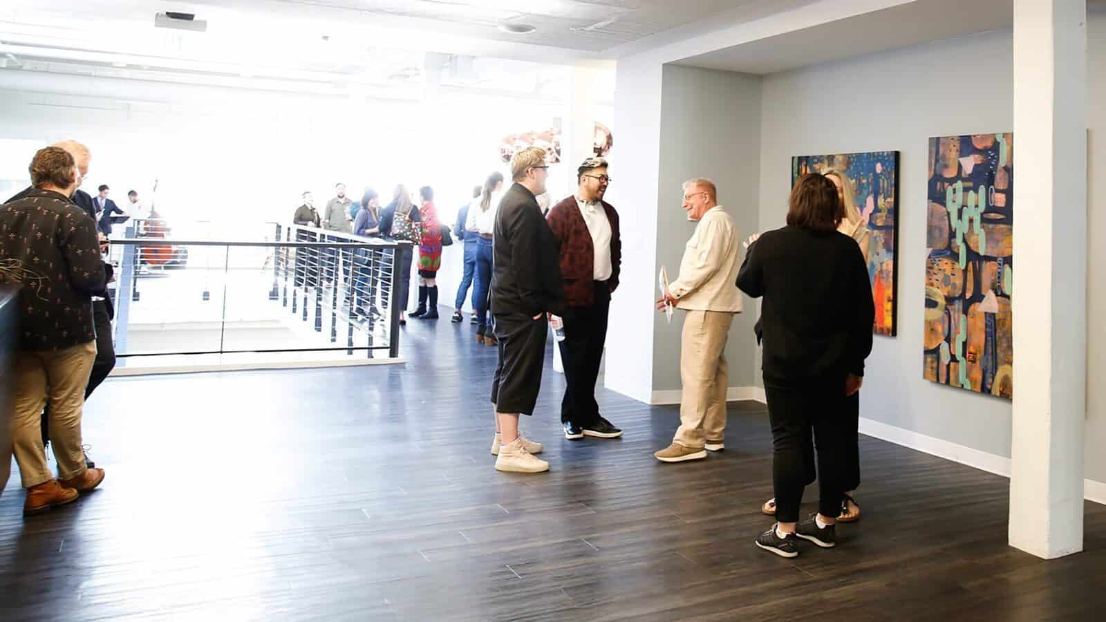 People converse and view abstract paintings in a modern gallery space with dark wood floors and white walls, highlighting the impact of Seattle Arts Funding; a group stands near a railing in the background.