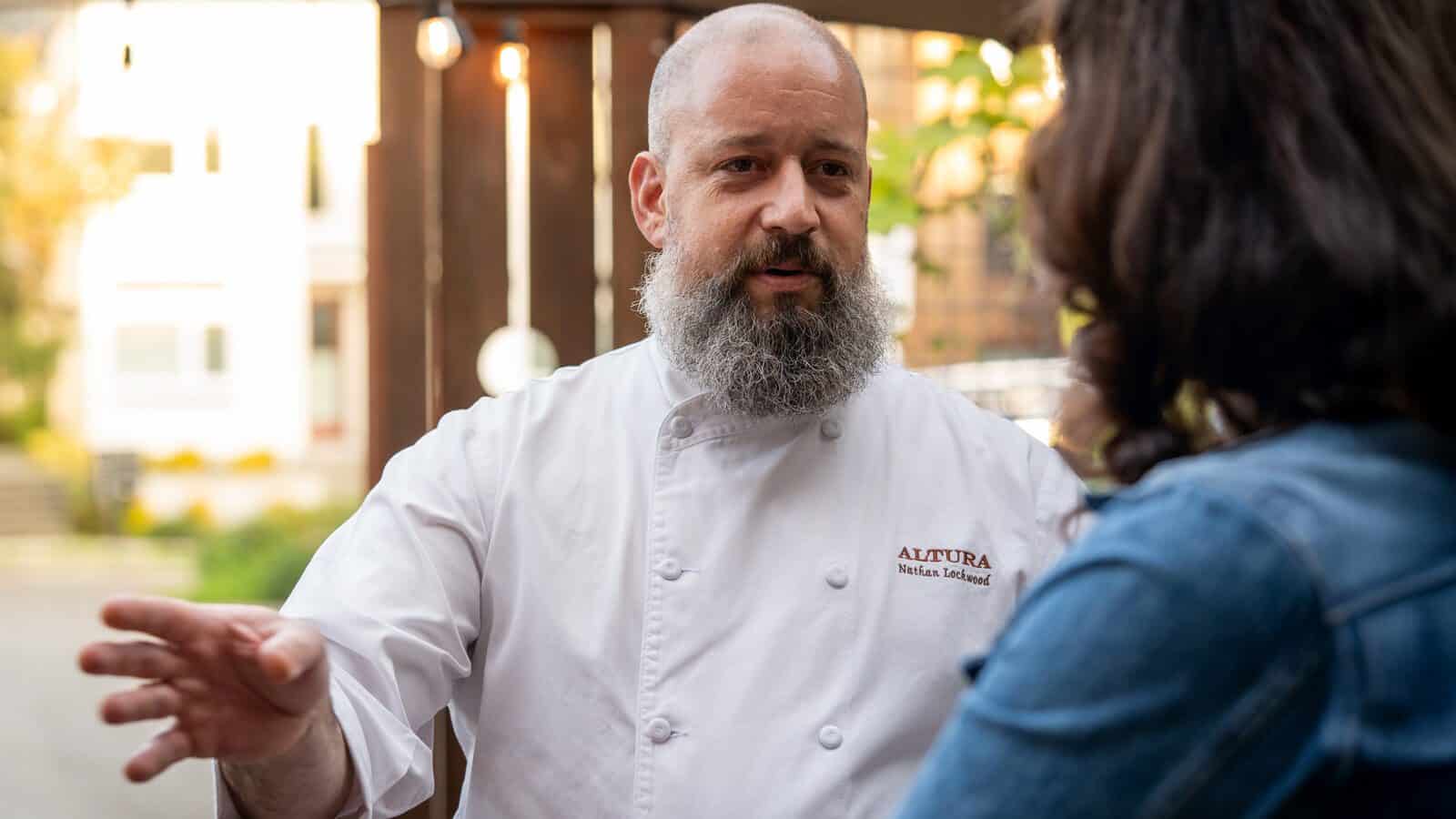 A chef in a white coat with "ALTURA" and a name embroidered on it speaks to a person in a denim jacket outdoors, inviting them to enjoy a summer visit at the restaurant.