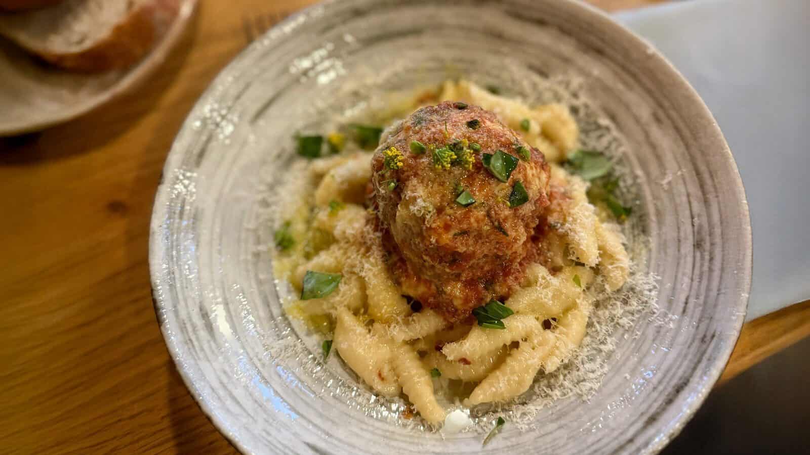 A round meatball sits on a bed of pasta, garnished with grated cheese and fresh herbs on a ceramic plate—perfect for a summer meal. Visit Carrello to enjoy this classic Italian comfort food.