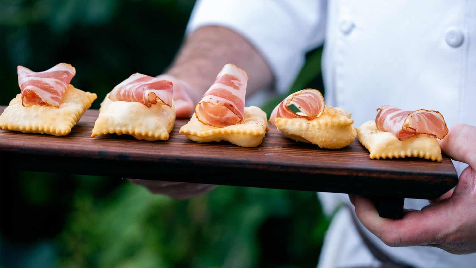 A person in a white chef’s coat holds a wooden board with five crispy crackers, each topped with cured meat—perfect for summer dining at Carrello.