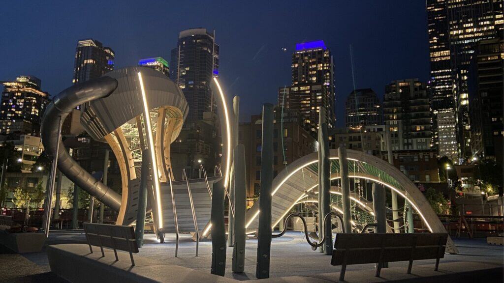 A modern playground with illuminated climbing structures and slides, set against a backdrop of tall city buildings at night.