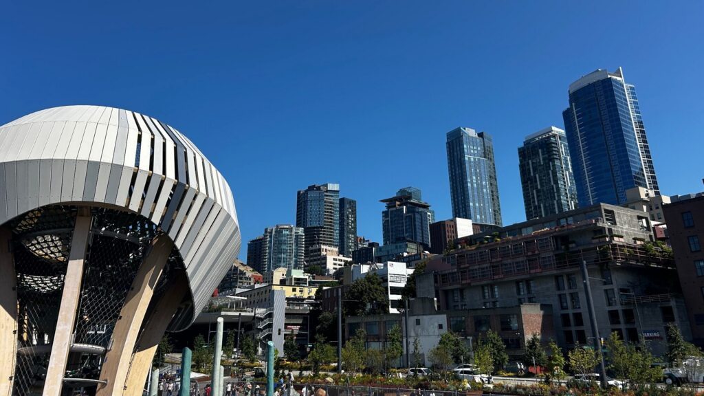 A modern cityscape featuring tall glass buildings under a clear blue sky, with a large, round, wooden playground structure in the foreground.