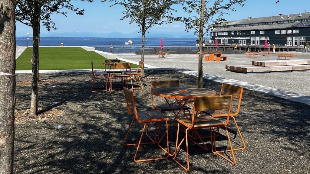 Outdoor seating area with orange tables and chairs under trees, near a waterfront with a building, grassy area, and benches visible in the background.