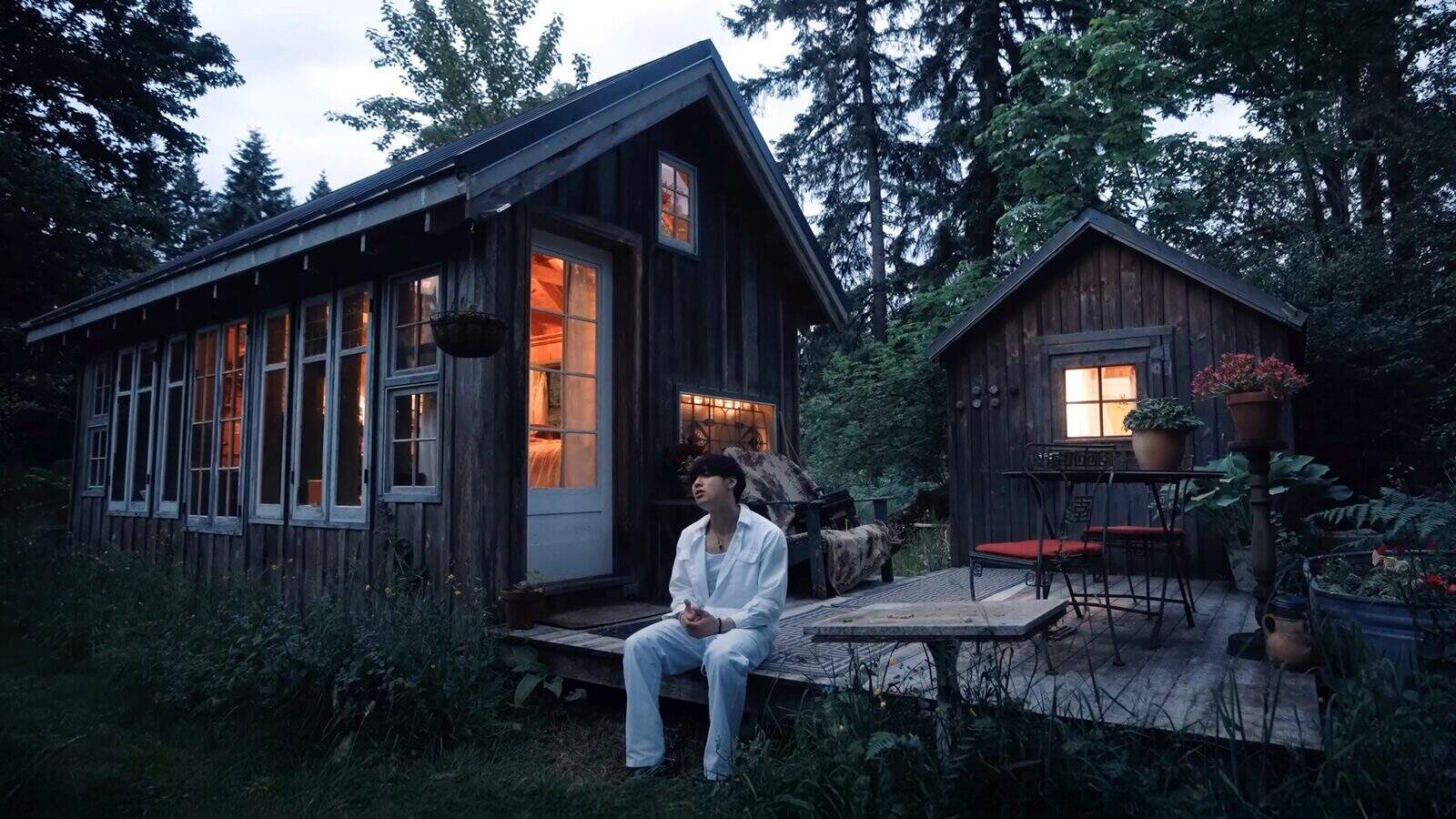 A person in white clothes sits on the porch of a rustic wooden cabin at dusk, with warm light glowing from the windows and surrounded by trees—a romantic scene inspired by a new music video from Johnny Huynh.
