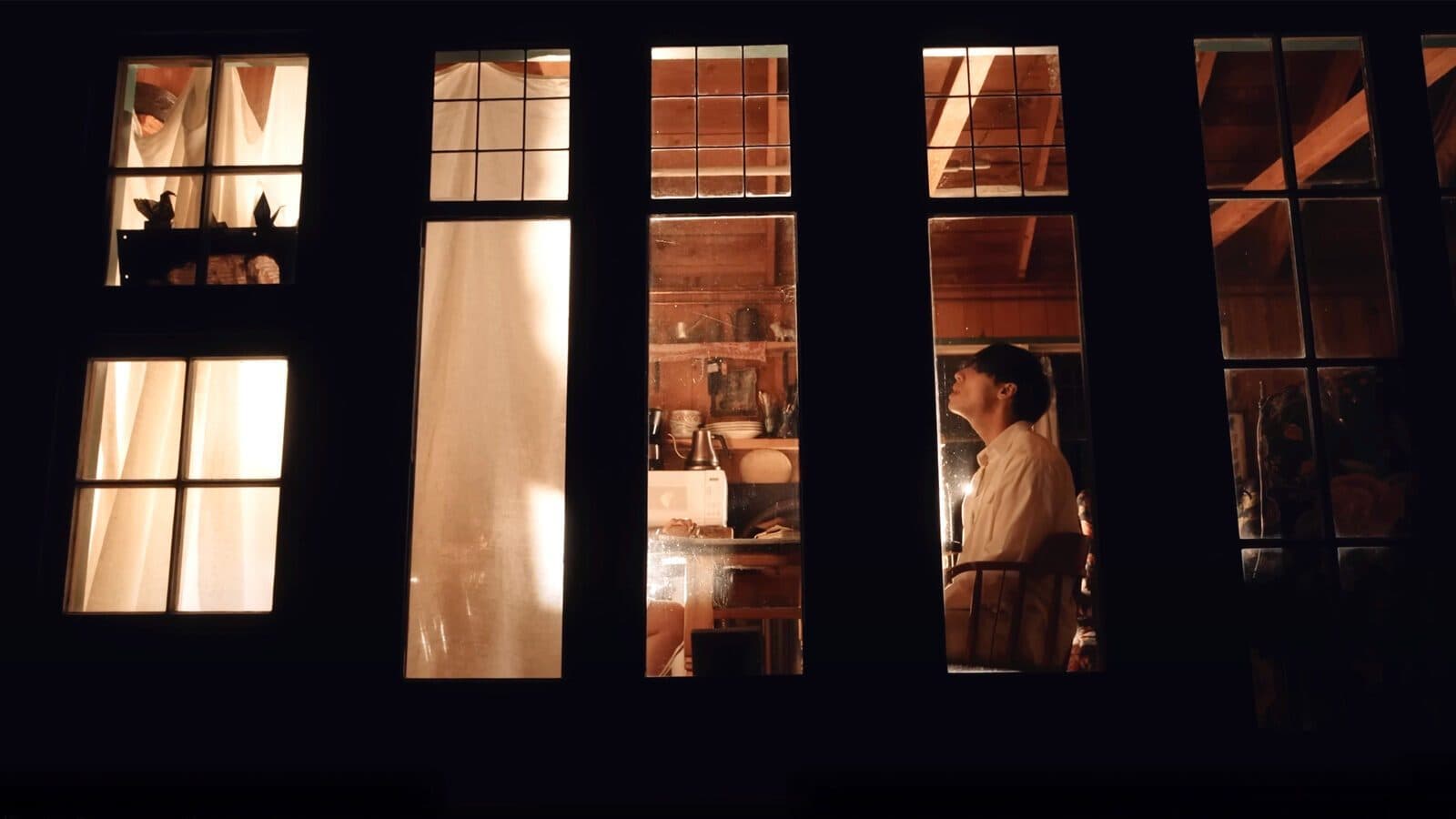 A moody and romantic nighttime view through multi-paned windows reveals two people in separate rooms—one behind a curtain, the other at a table—captured like a scene from Johnny Huynh's new music video.