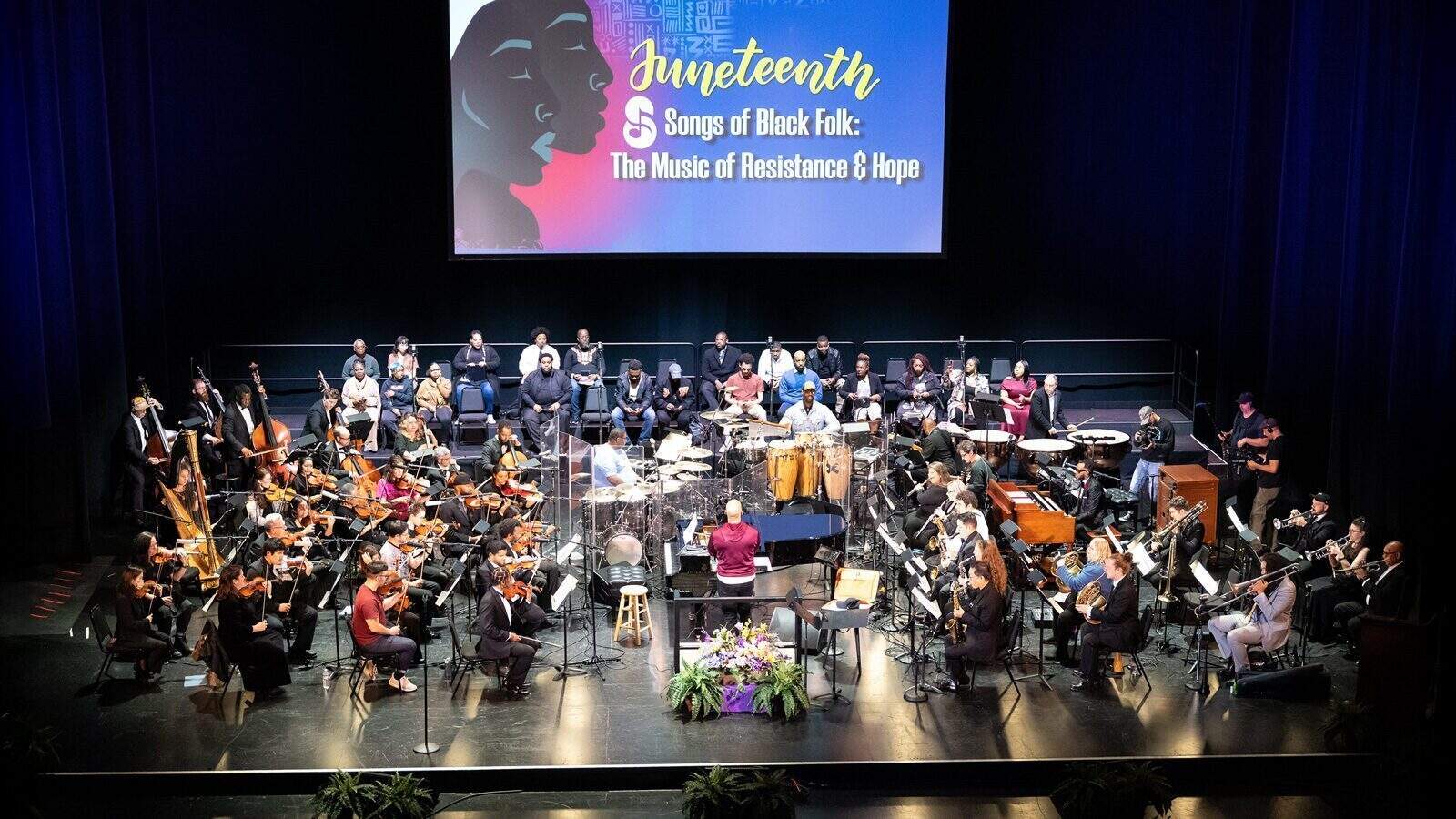 An orchestra and choir perform on stage beneath a screen displaying “Juneteenth: Songs of Black Folk – The Music of Resistance & Hope,” marking its Tribeca Debut.