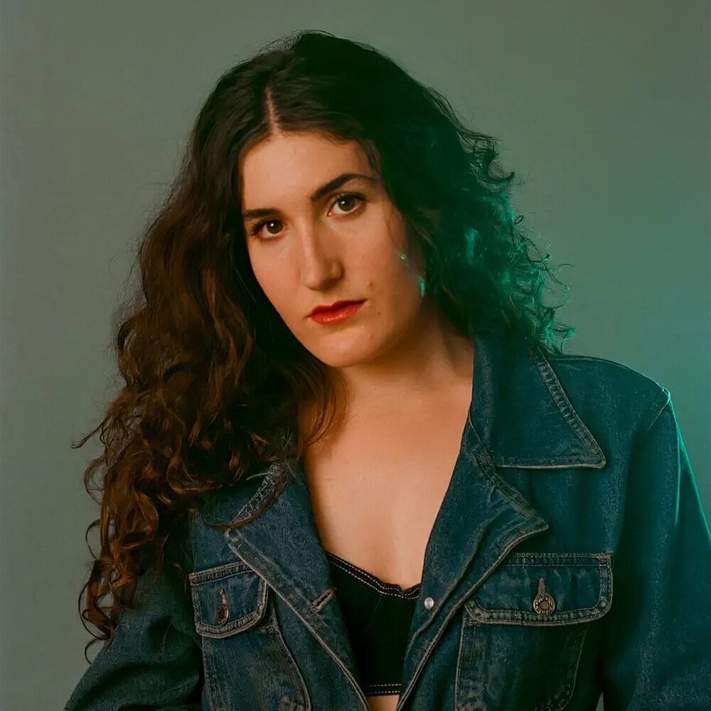 A woman with long curly hair wearing a denim jacket and a black top poses in front of a plain background—a must list look for 07-24-2025.