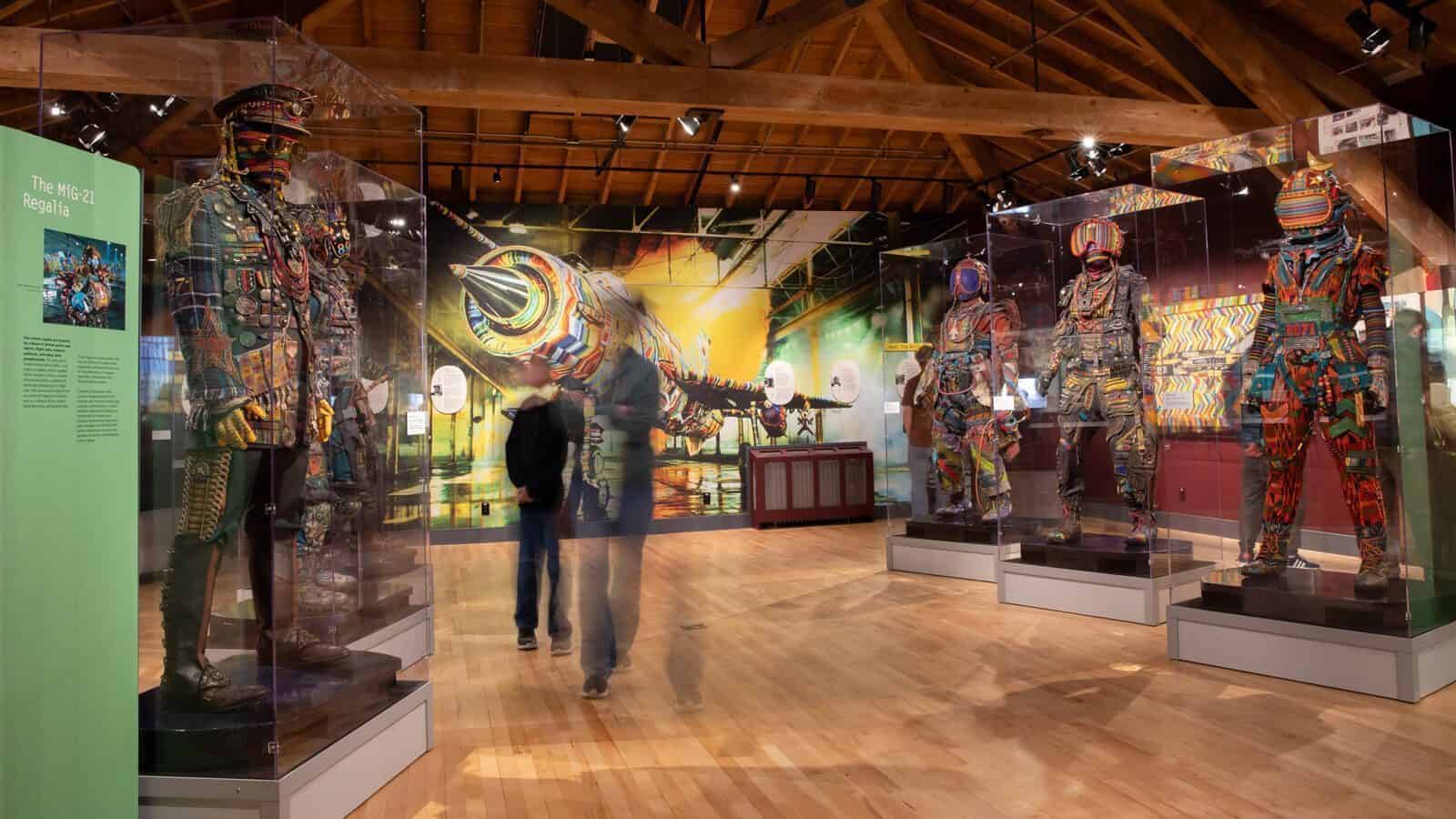 Exhibit room with four colorful humanoid sculptures in glass cases, two visitors walking by, and a vibrant mural of a jet in the background, blending art and war themes.