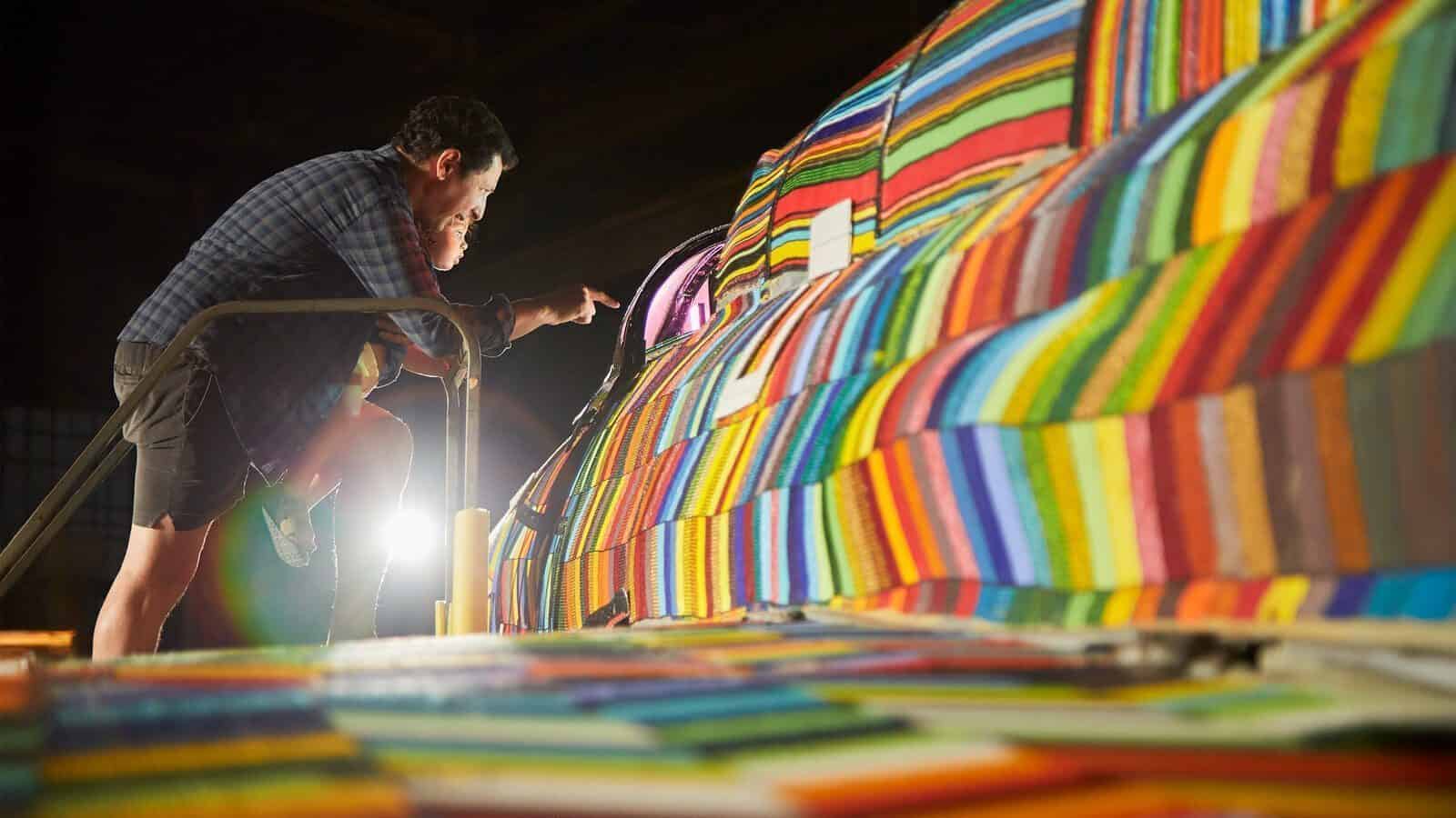 A person inspects a large object covered in colorful, striped fabric panels inside a well-lit indoor space, reflecting the theme of turning war into art.
