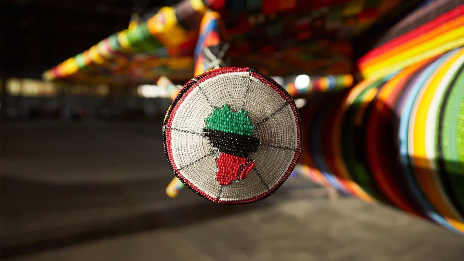 A close-up of a round, beaded ornament featuring a map of Africa, suspended in front of a colorful, yarn-wrapped art structure.