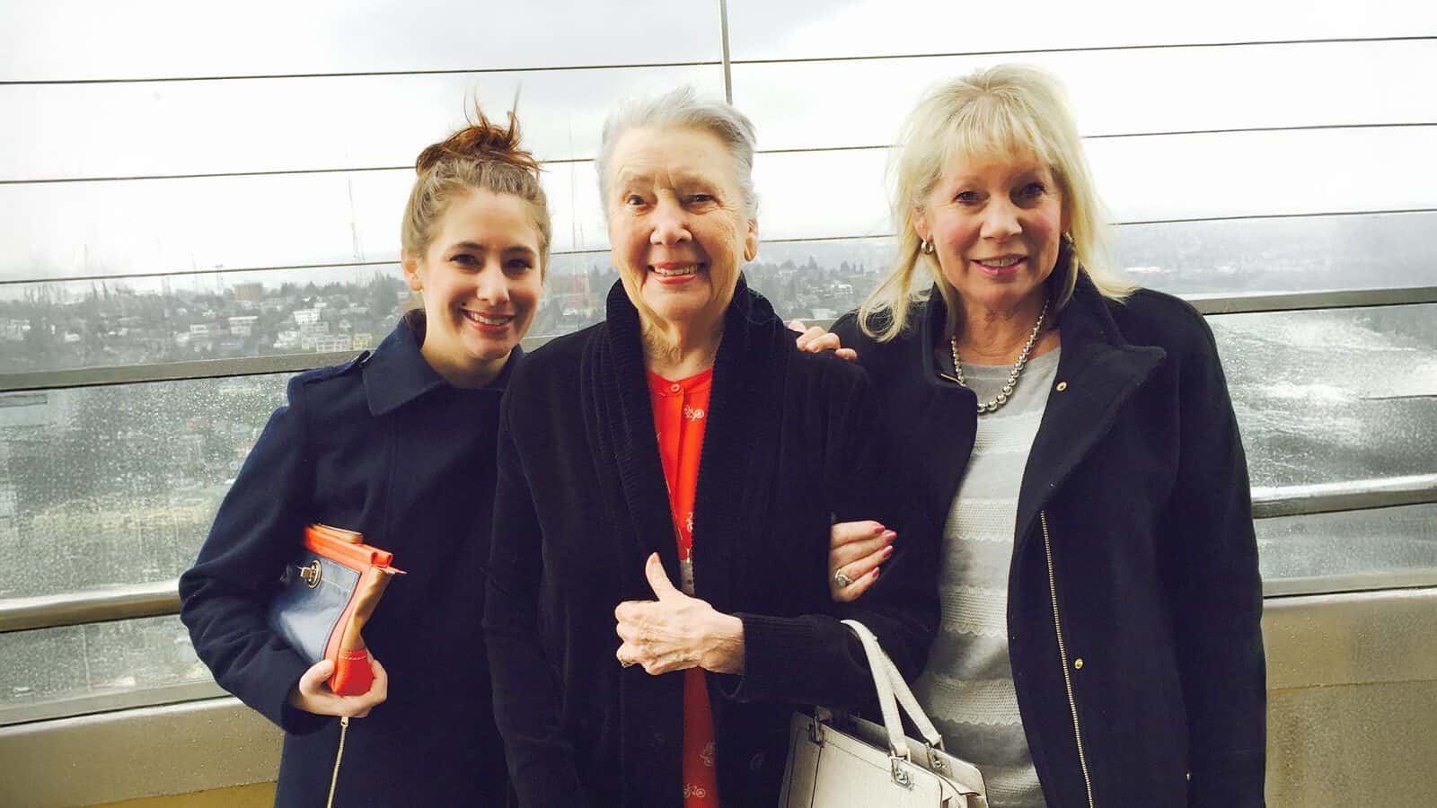 Three women stand together smiling, dressed in jackets, with a rainy cityscape visible through the windows behind them—a moment captured for Seattle Magazine by its Editor in Chief.