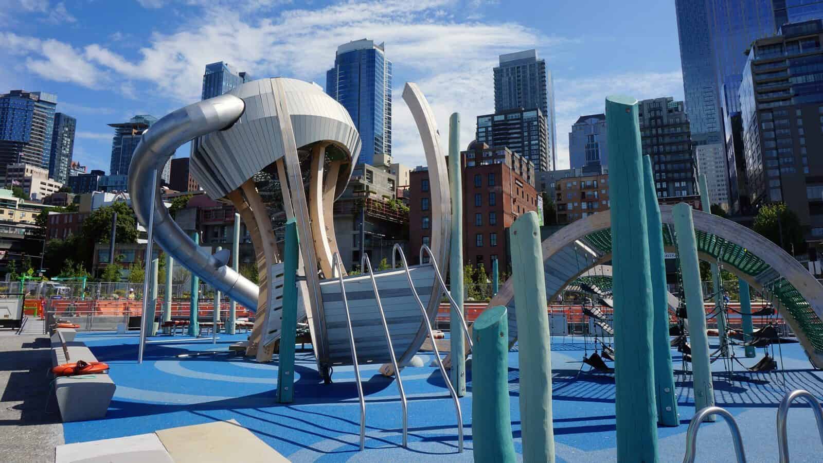 Pier58-Playzone_1600x900 Modern urban playground with climbing structures and slides offers endless playtime fun, set against a backdrop of tall buildings and a blue sky near the lively pier.
