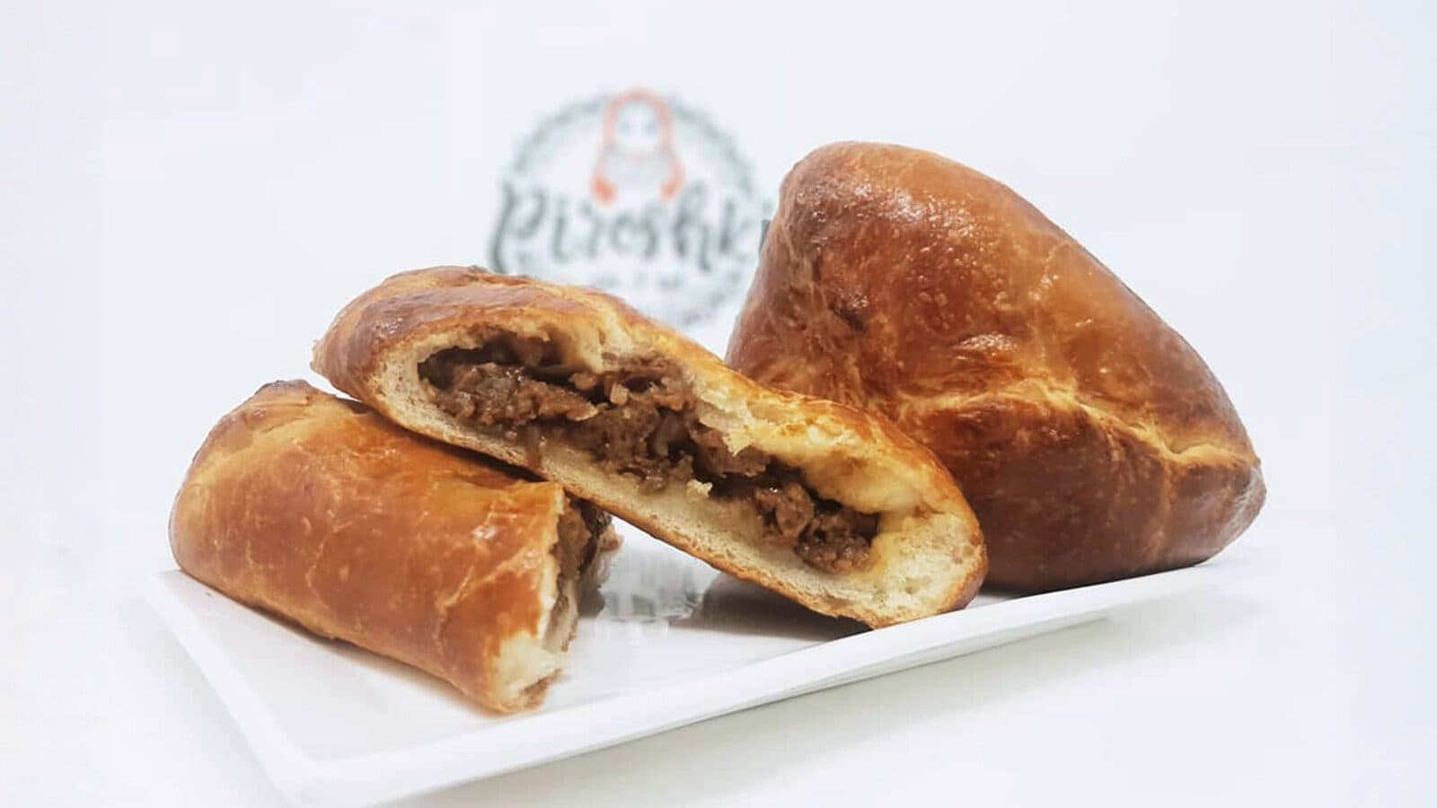 A baked pastry stuffed with seasoned ground meat, perfect for seasonal eating, shown whole and halved to reveal the filling, placed on a white plate with a blurred logo in the background.