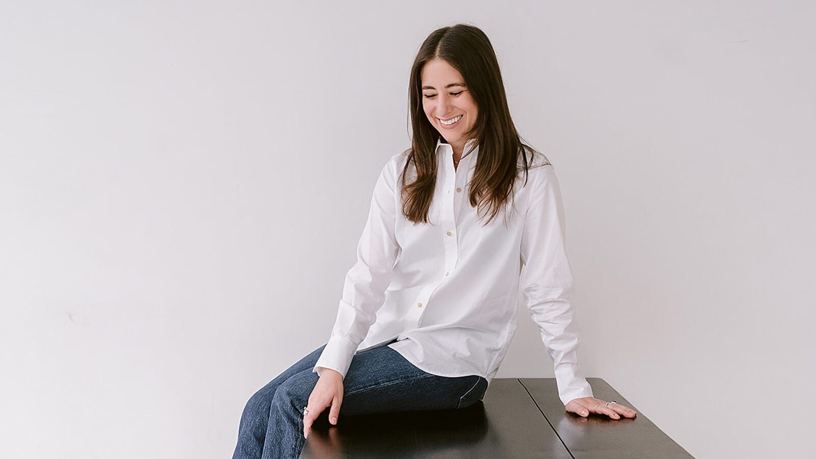 A woman with long brown hair, wearing a white button-up shirt and blue jeans, sits on the edge of a dark table against a plain white background, smiling and looking down.