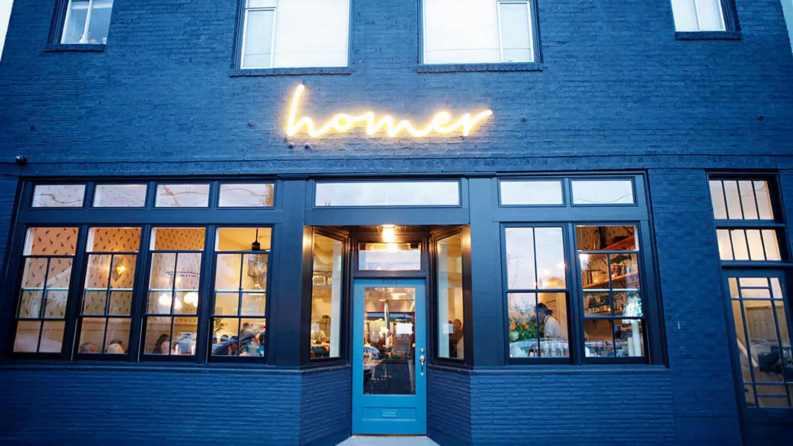 RestaurantHomer_1600x900 Exterior of a dark blue brick restaurant building with large windows and a lit sign above the door reading “homer”; indoor lighting and diners visible inside, welcoming guests for the Homer Reopening featured in Restaurant Roundup.