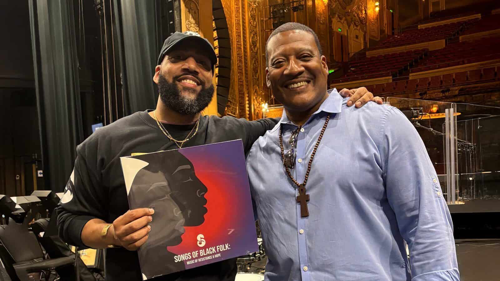 Two men stand together on a theater stage; one holds the vinyl record "Songs of Black Folk" with abstract artwork, both smiling at the camera after its Tribeca Debut.