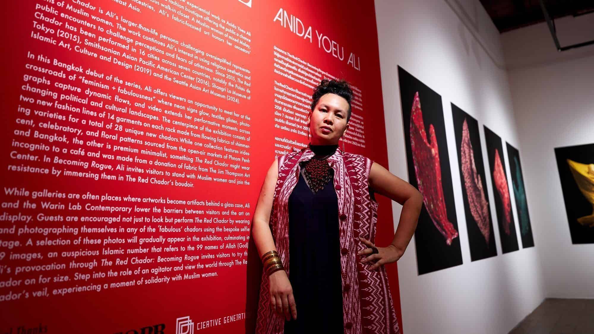 A woman stands confidently in front of a red exhibit wall featuring information about Anida Yoeu Ali, with framed artwork displayed on the adjacent wall.