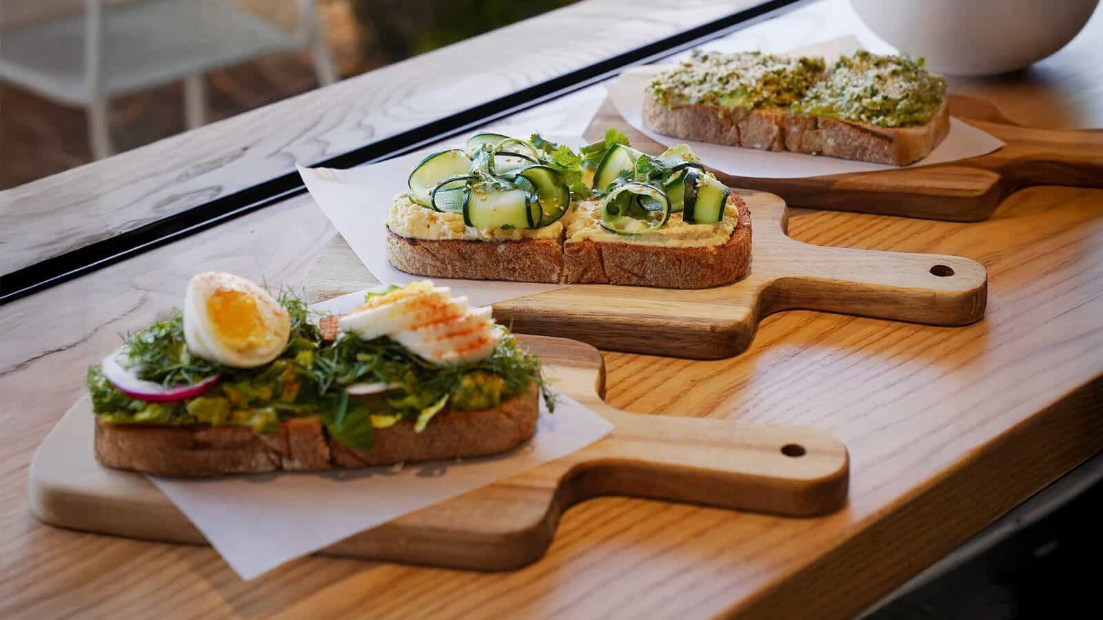 Sano-Bowl-Toast_1600x900 Three open-faced sandwiches with various toppings, including sliced egg, vegetables, and herbs, are arranged on wooden boards at Sano's Eastside locations.