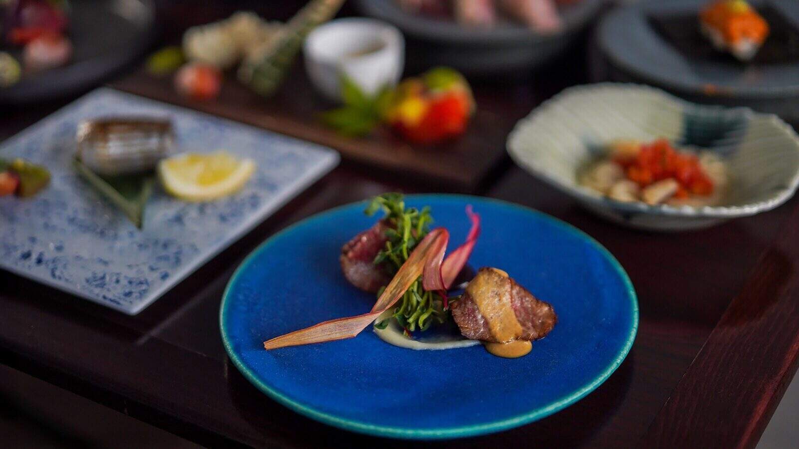 ShomonKappoSushi_1600x900 A blue plate with seared meat, sauce, and garnish is in focus; other assorted dishes are blurred in the background on a dark table.