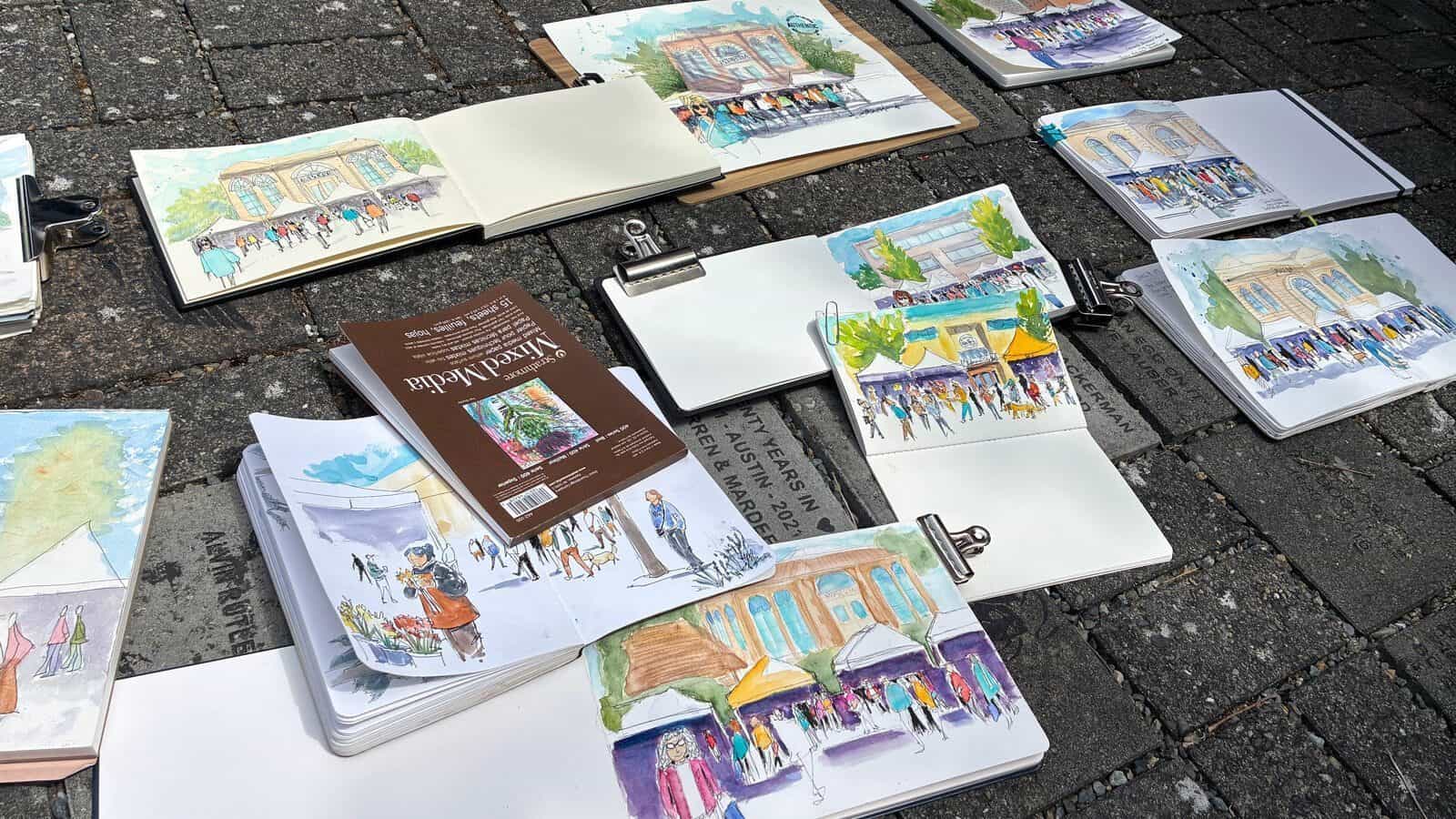 Several sketchbooks and drawings of street scenes and buildings are laid out on a stone pavement, accompanied by drawing supplies and a brown notebook—perfect tools for art observation and visual analysis.