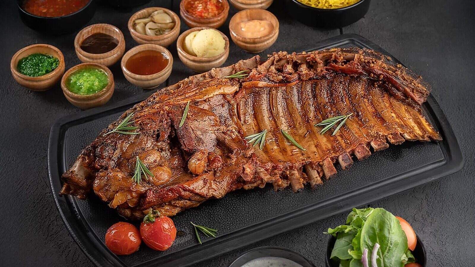 SmokeMeat-MeatMootRestaurant_1600x900 A large cooked rack of ribs from PCC Corner Market is garnished with rosemary and served on a black tray, accompanied by cherry tomatoes, sauces in small bowls, and a side salad—a true high-end halal feast.