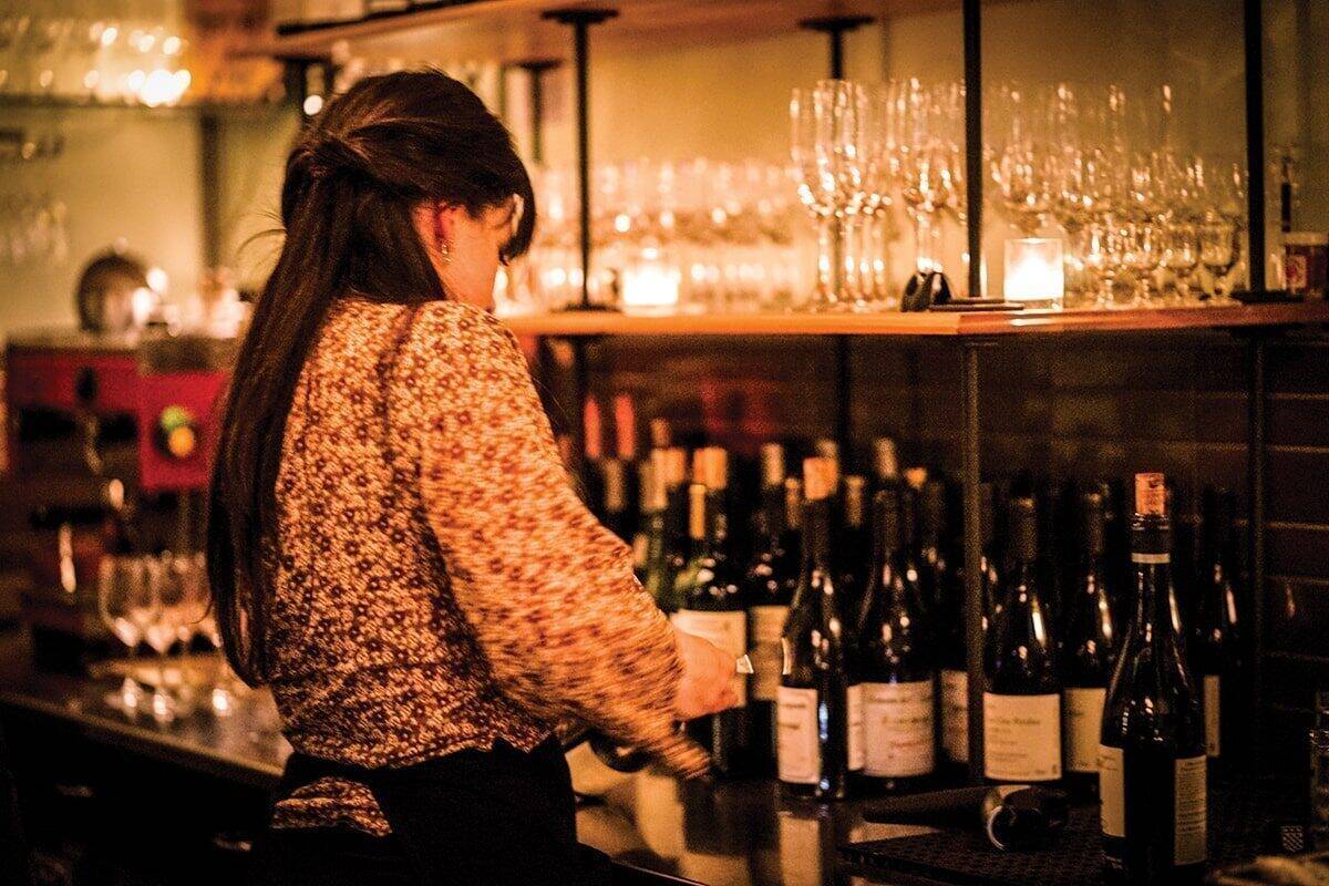 A bartender with long hair tied back stands behind a bar, surrounded by wine bottles and glassware, preparing drinks in a dimly lit setting reminiscent of summer in Seattle.