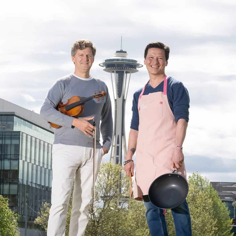 Two men stand outdoors in front of the Space Needle; one holds a violin and bow, the other wears an apron and holds a frying pan and rolling pin. Don’t miss this Must List moment—save the date: 07-24-2025!.