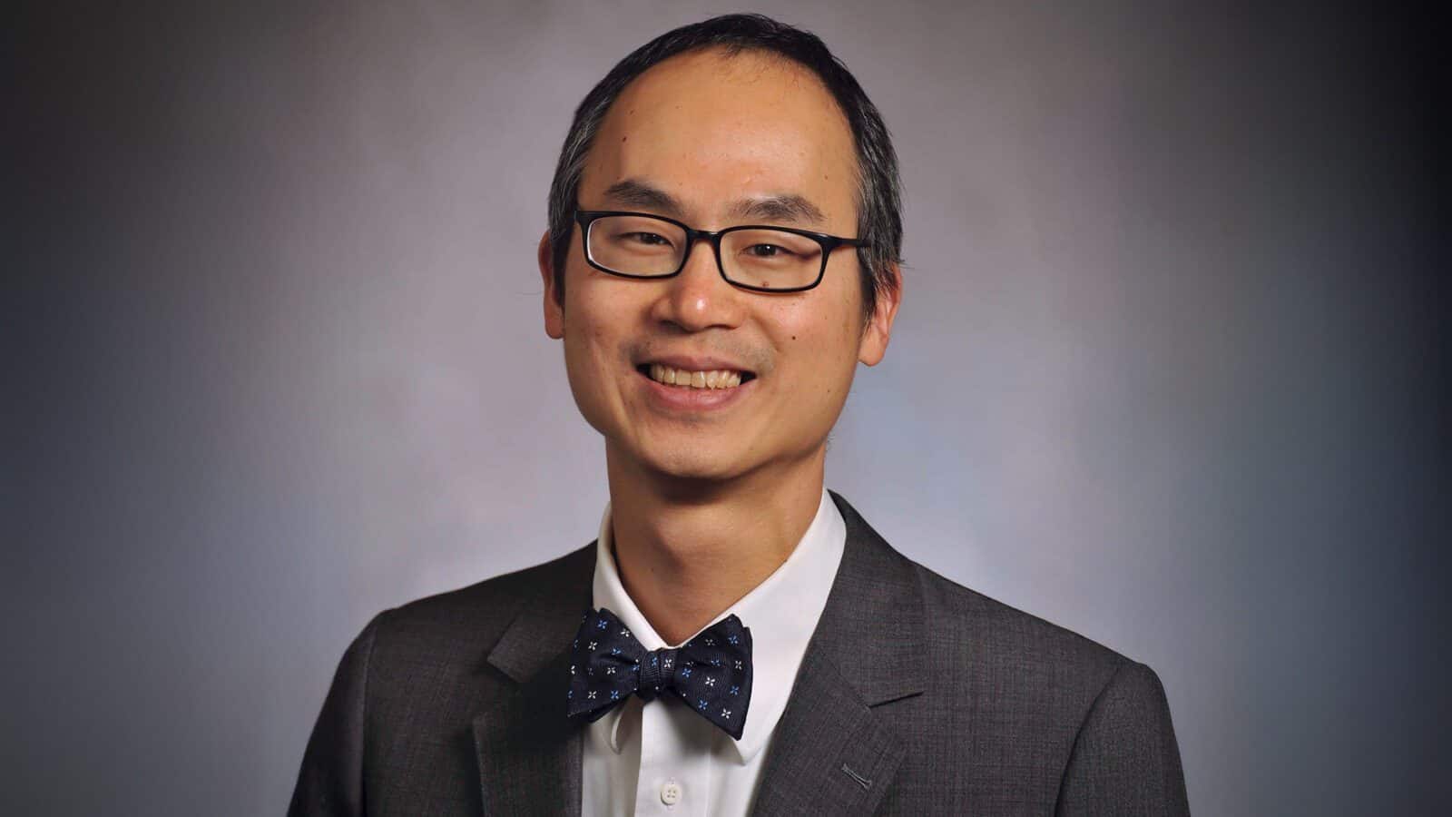 Dr. Tobias Lee, M.D., wearing glasses, a dark suit, a white shirt, and a polka dot bow tie, smiles in front of a plain background.