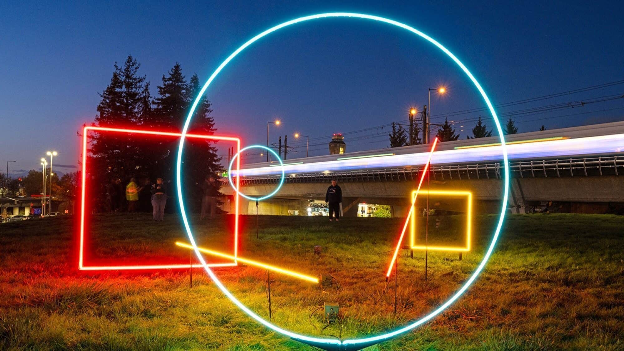 Glowing neon shapes—circle, square, and lines—are displayed outdoors at dusk, with people, trees, and a bridge with a moving train in the background.