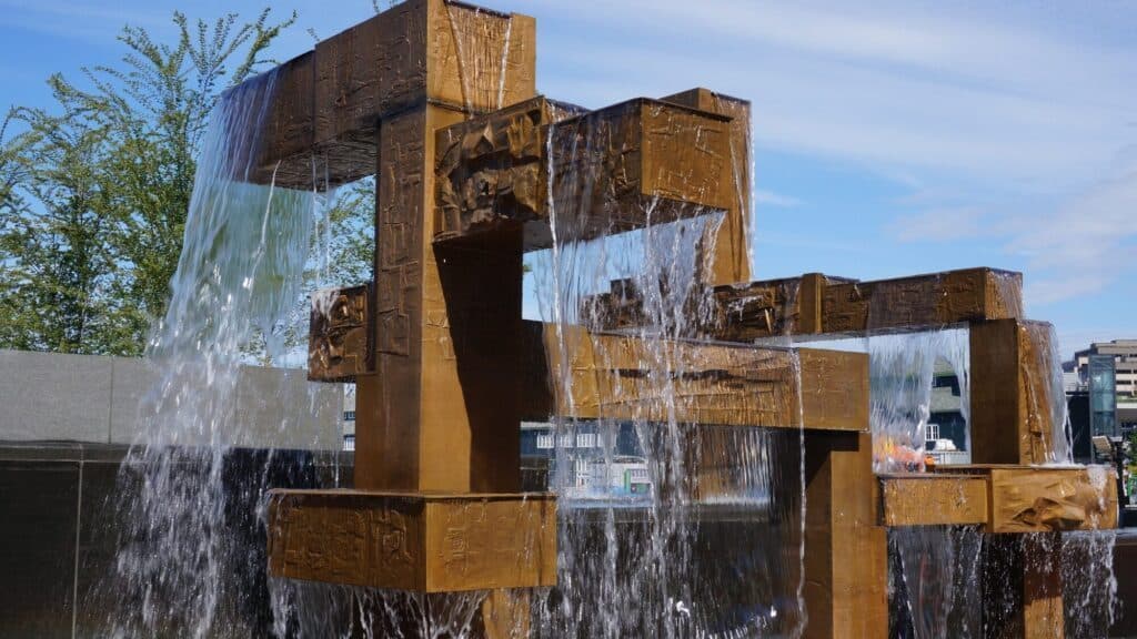 Bronze-colored geometric metal fountain structure with water cascading over its rectangular shapes, set outdoors against a blue sky and some greenery.