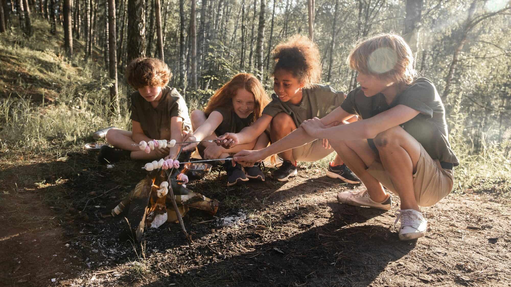 Four children crouch by a small campfire, roasting marshmallows on sticks in a forested area during daylight.
