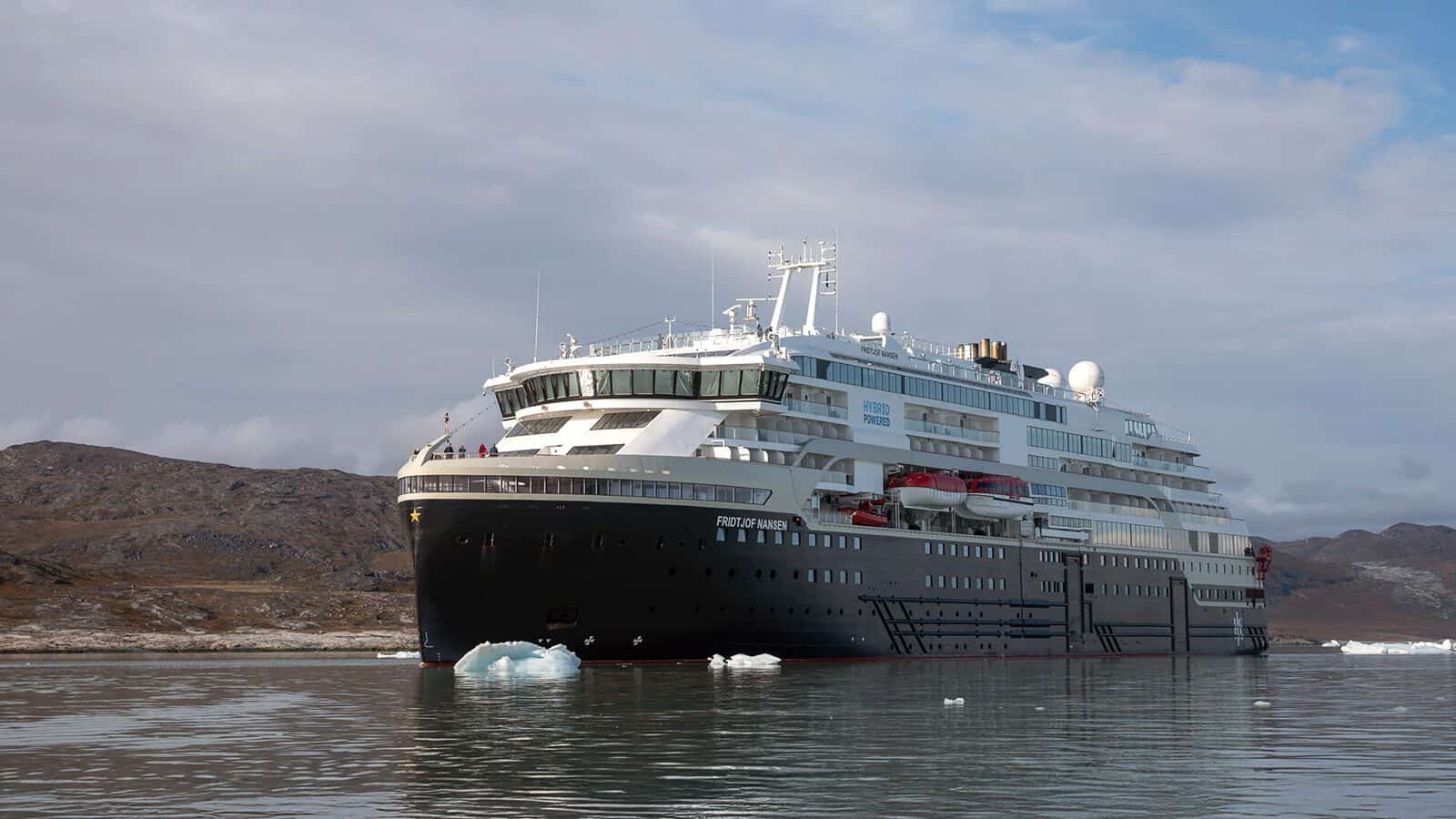 A large modern cruise ship is anchored in calm Arctic waters, surrounded by floating icebergs and rocky hills—an unforgettable scene on a Greenland cruise.