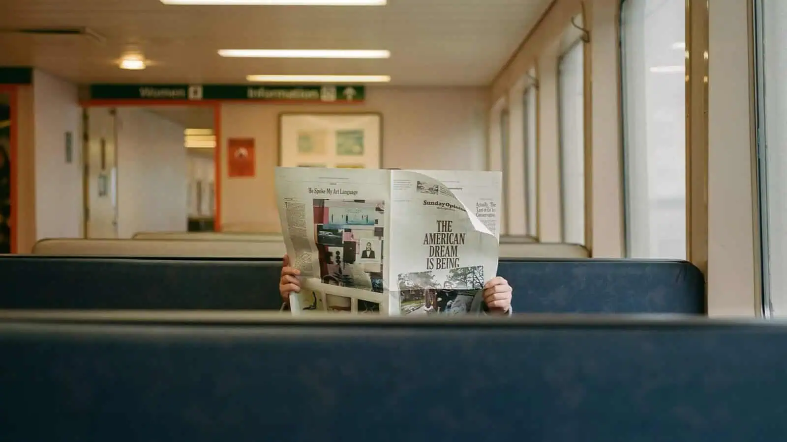 A person sits alone on a Seattle ferries bench, face hidden behind a newspaper titled "The American Dream is Being...," in a candid moment captured by Kristopher Shinn using 35mm photography.