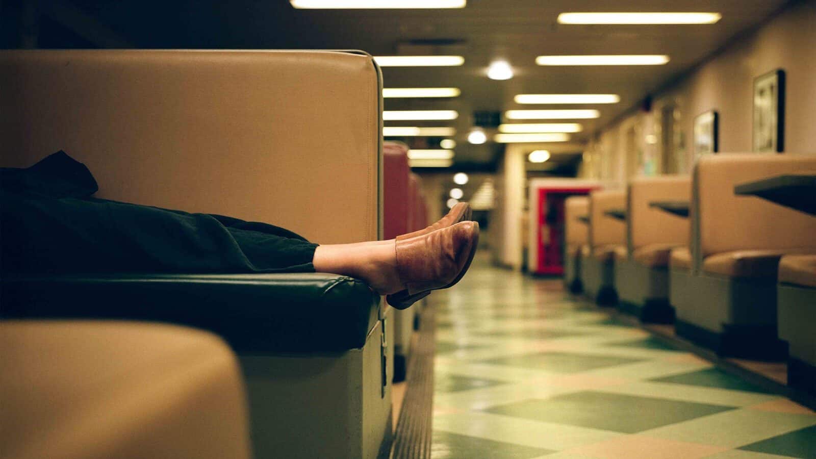 A person lounges in a booth at an empty diner, with legs stretched out and feet resting on the opposite seat. The brightly lit space captures a quiet moment reminiscent of Kristopher Shinn's 35mm photography style.