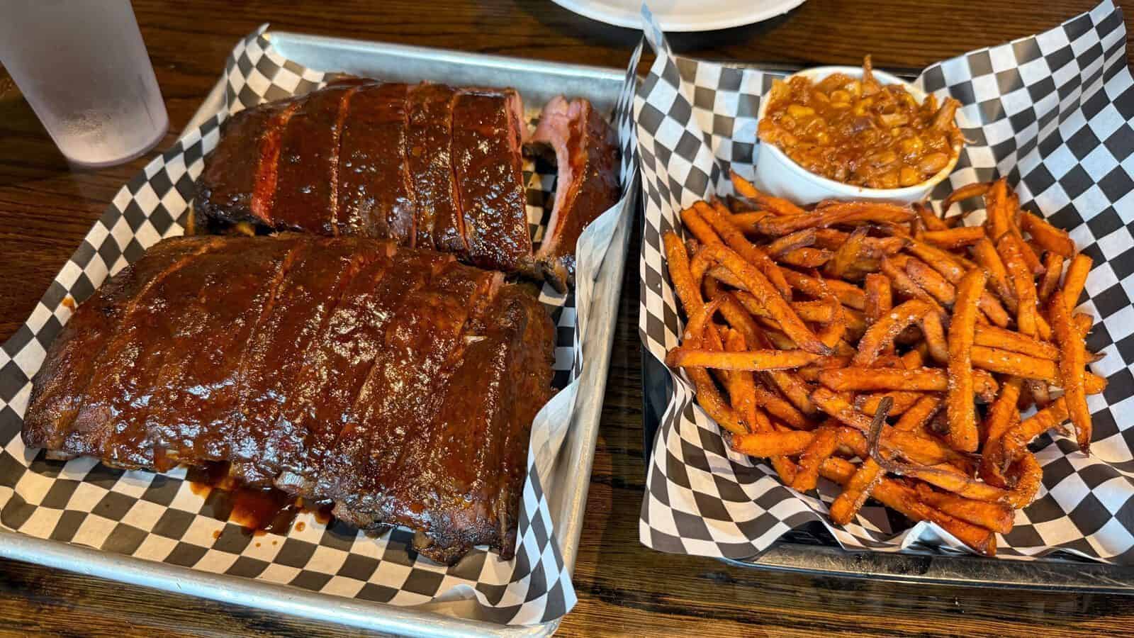 Two racks of glazed BBQ ribs from Brileys BBQ rest on a tray with sweet potato fries and baked beans, all served atop checkered paper—a true taste of Counter Culture.