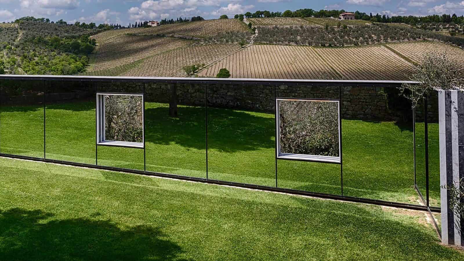 A mirrored fence with two window-like openings reflects the grassy lawn and surrounding landscape of fields and hills, creating a striking Art Among the Vines installation.