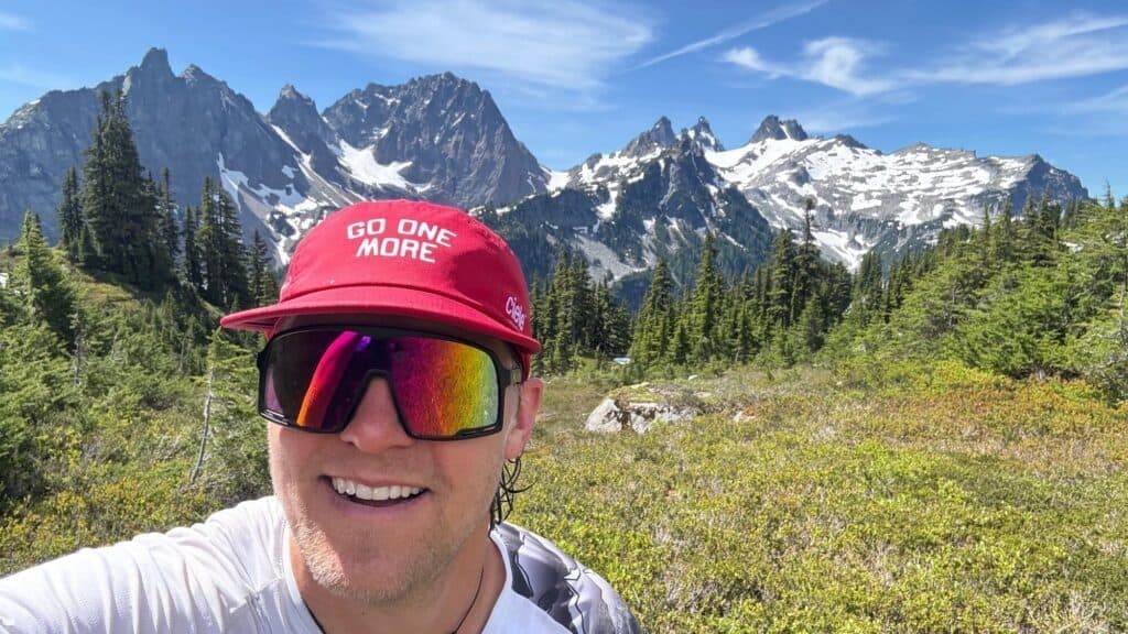Person wearing a red hat and reflective sunglasses smiles in front of snow-capped mountains and evergreen trees on a sunny day.