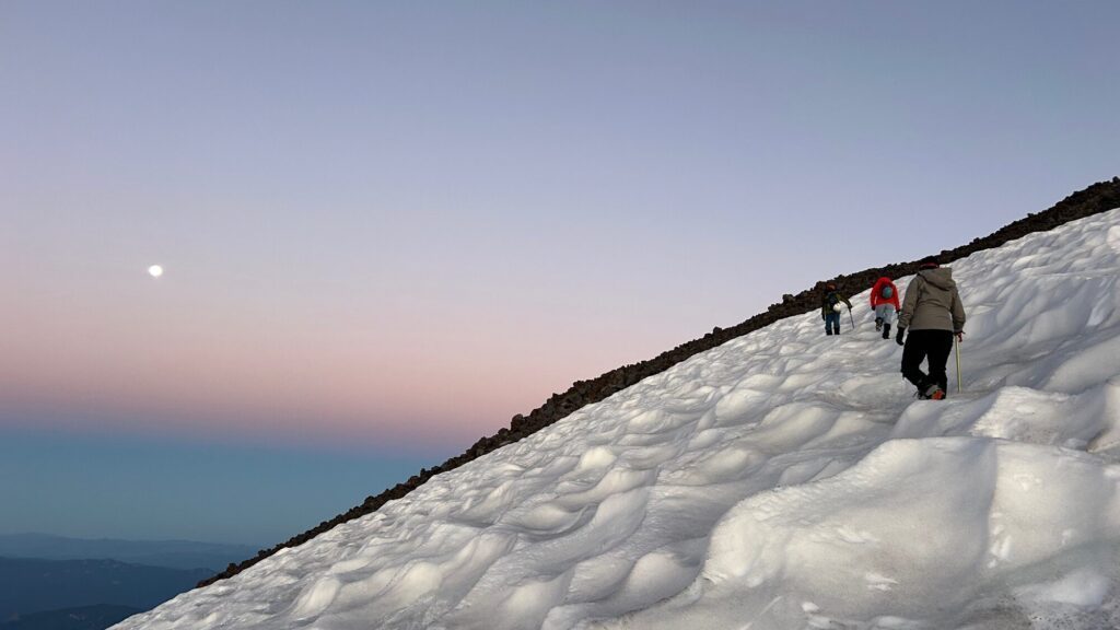 A group of people hike up a snowy, sloped mountain at dusk with the moon visible in the sky.