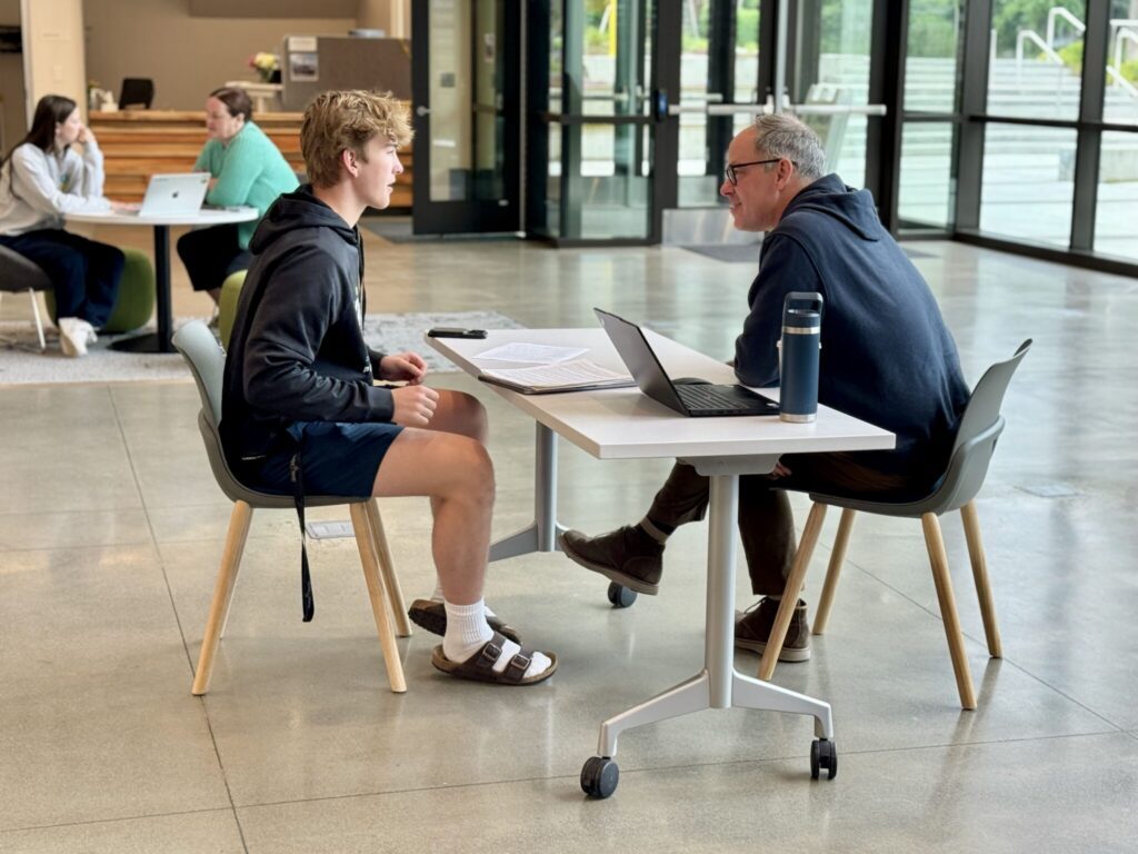 A student and an adult sit across from each other at a table with laptops and papers, discussing college admission in a bright, modern common area at UPrep private school. Other people are seated and talking in the background.