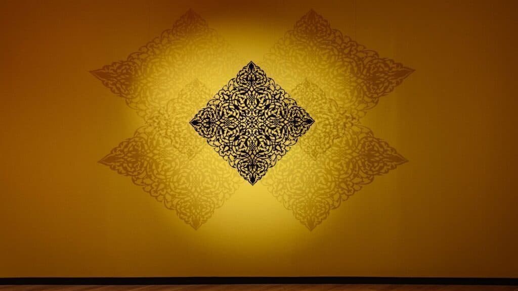 A black ornamental art piece is mounted on a yellow wall, casting symmetrical decorative shadows in a diamond pattern.