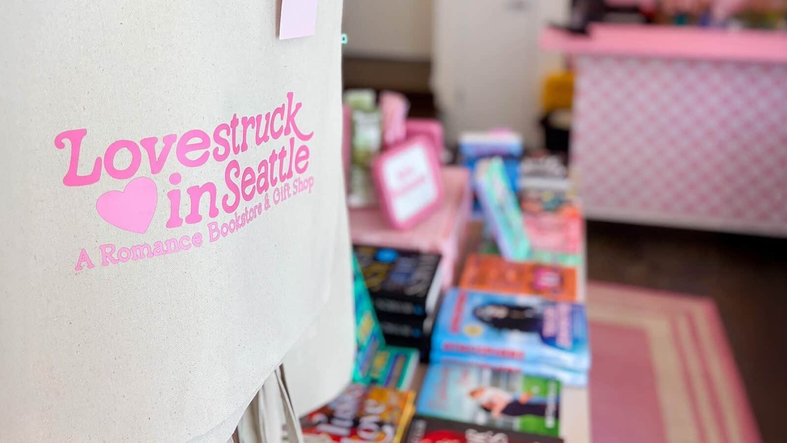 Tote bag with "Lovestruck in Seattle: A Romance Bookstore & Gift Shop" logo in focus; blurred background shows colorful books and a pink counter—perfect for carrying your next love story from the bookstore.
