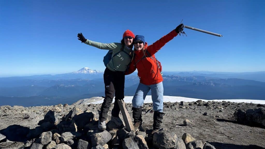 Two people in hiking gear stand on a rocky mountain summit with arms outstretched, smiling, with a snowy peak and clear blue sky in the background.