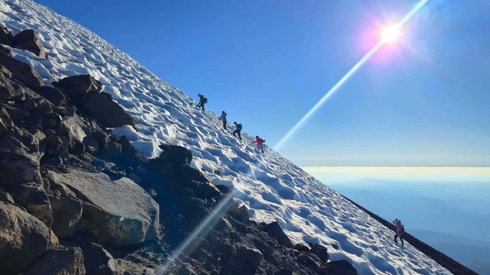 Climbers ascend a steep, snow-covered mountain slope under bright sunlight, with clear blue sky and scattered rocks visible.