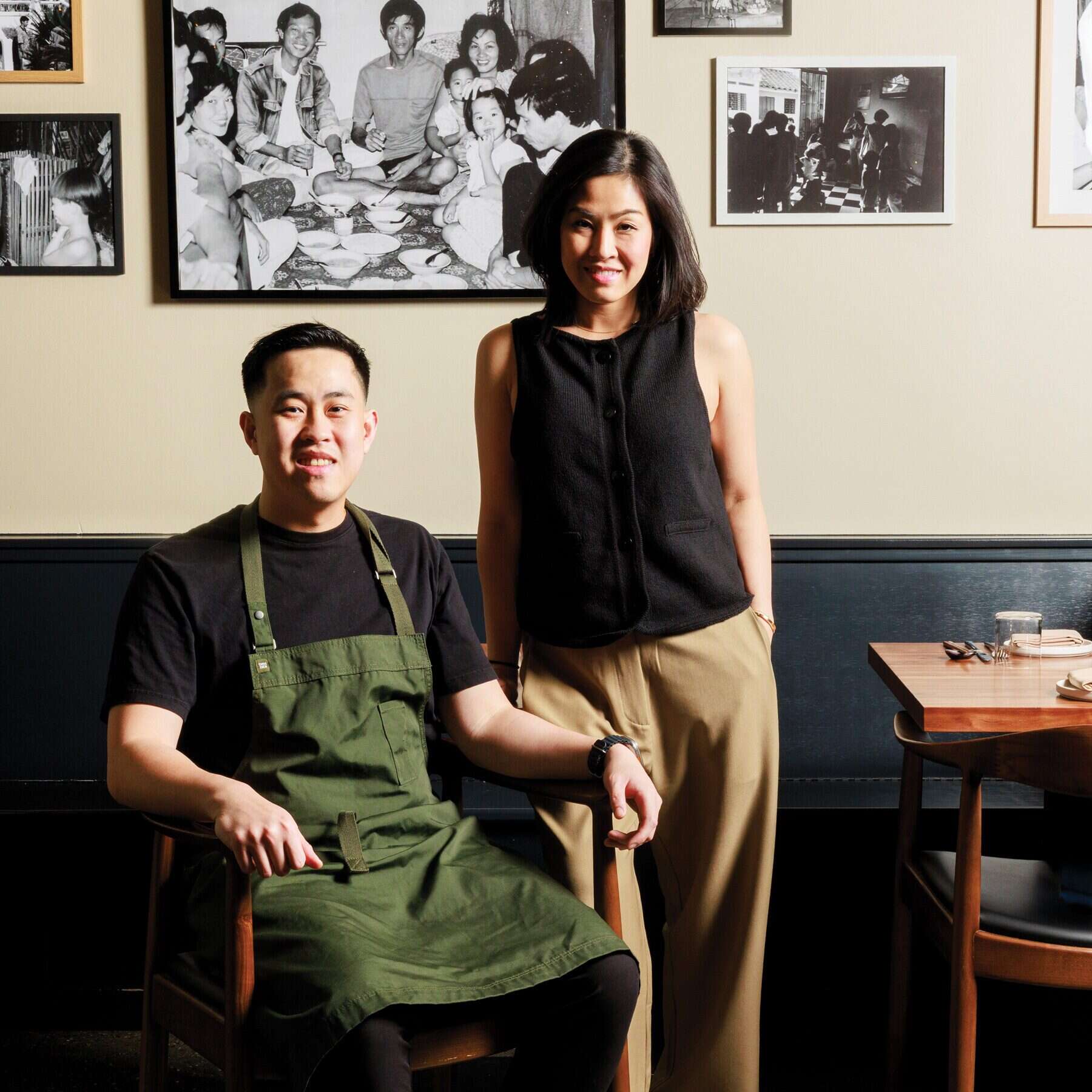 A man in a green apron sits next to a woman standing by a dining table at Ramie, a Capitol Hill restaurant known for its Vietnamese cuisine, with black-and-white photos on the wall behind them.